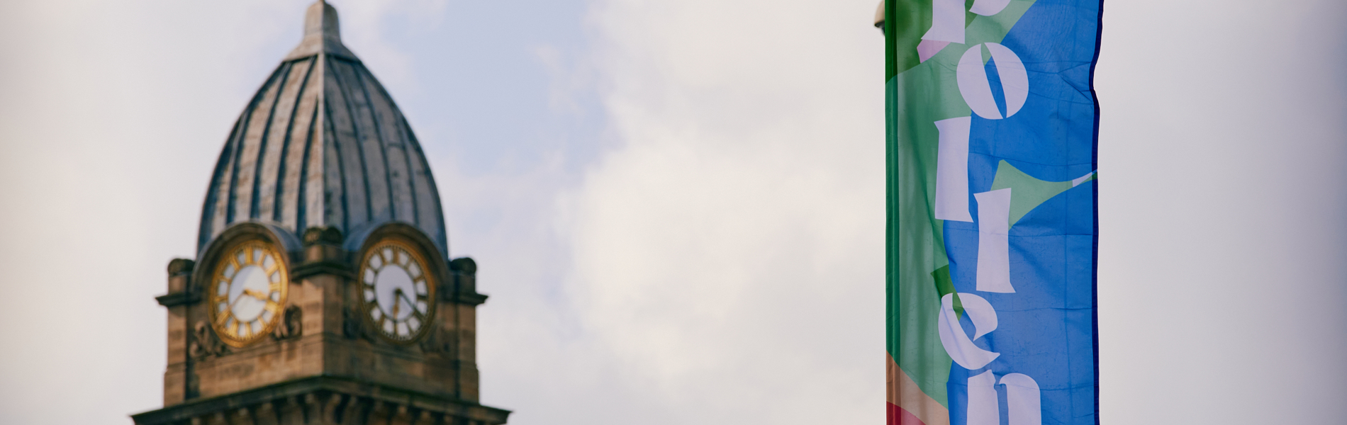In the foreground is a colourful flag that has the word 'Pollen' on it, in the background is a clock tower.