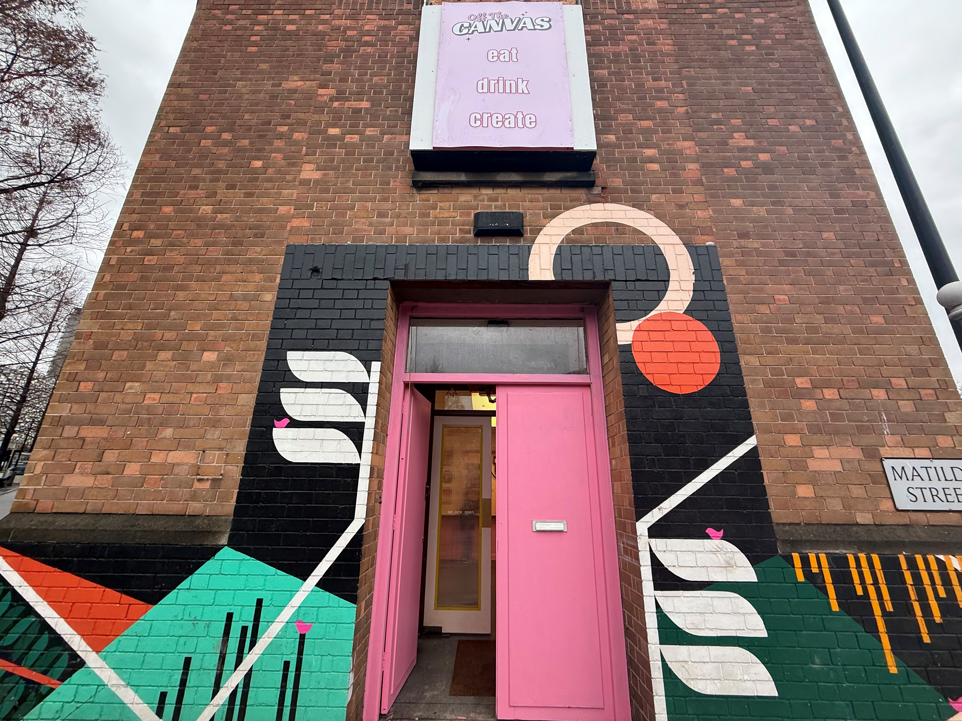 The exterior of a brick building with a pink doorway framed by bold, geometric street art and a sign above reading “eat drink create.”