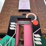 The exterior of a brick building with a pink doorway framed by bold, geometric street art and a sign above reading “eat drink create.”