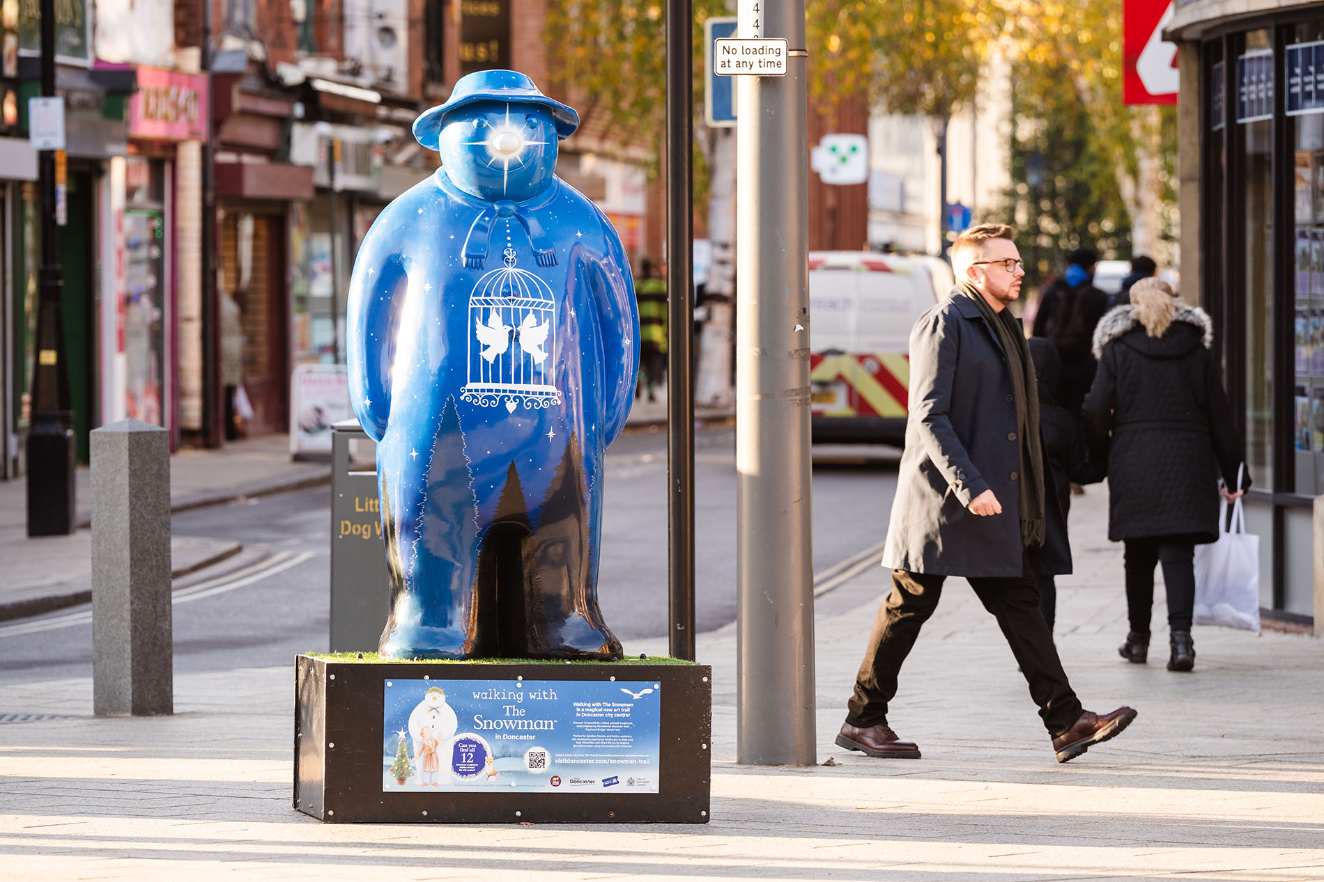 A large blue snowman sculpture decorated with white stars and an illustration of two figures inside a snow globe stands on a black platform in a pedestrian street. Surrounding the sculpture are shops, a street sign, and people walking along the pavement. The scene is bright and sunny, with autumn-coloured trees visible in the background.