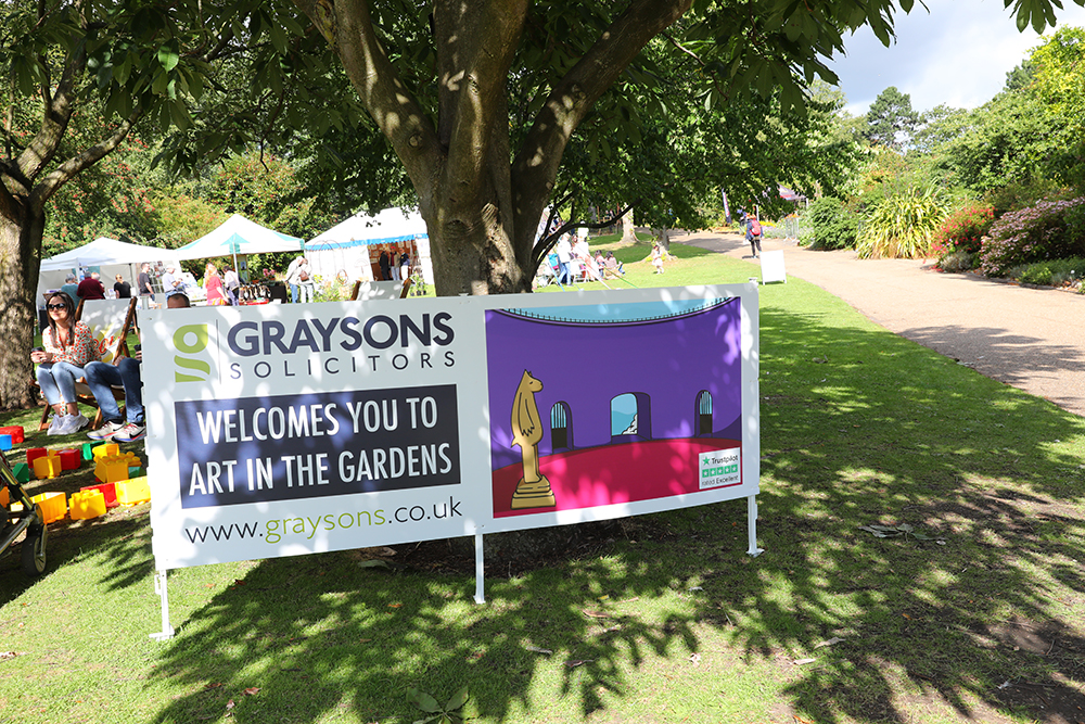 A large banner displayed outdoors under a tree reads “Graysons Solicitors welcomes you to Art in the Gardens” with the website www.graysons.co.uk. The banner features a colourful illustration of an art gallery with a sculpture and arched doorway. Behind it, people are seated near white tents on a grassy area, and a wide path leads through landscaped gardens with trees and shrubs in bright sunlight.