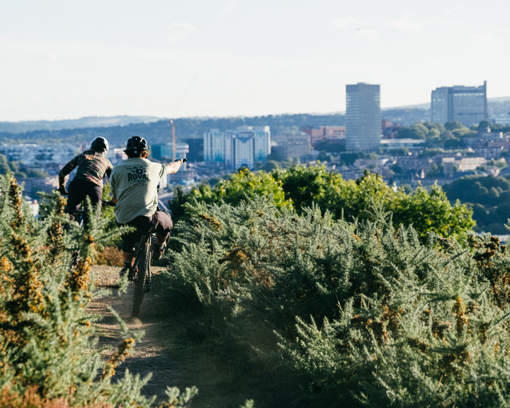 A man and a woman are mountain biking along a dirt track. In the distance you can see Sheffield city centre.