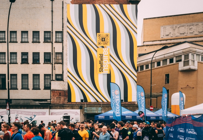 A massive banner on the side of the building shows the distinctive Sheffield Inspires branding as runners gather for Sheffield Half Marathon below it 