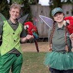 A man and a woman, dressed in green and wearing fairy wings, hold brightly coloured parrots at the Tropical Butterfly House Wildlife Conservation Park.