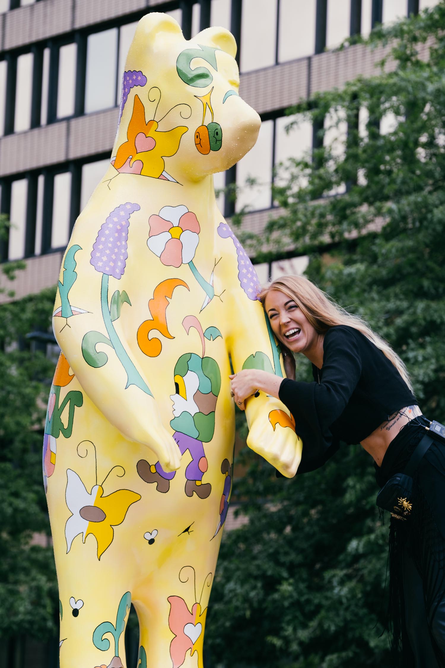 Large yellow bear sculpture decorated with colorful floral and abstract patterns, positioned outdoors near a modern building. A person is interacting with the sculpture, holding its arm