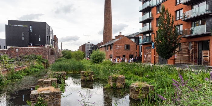 Building, a mixture of old and new, stand along the banks of a river. In the distance is a chimney stack rising up out of Kelham Island Museum.