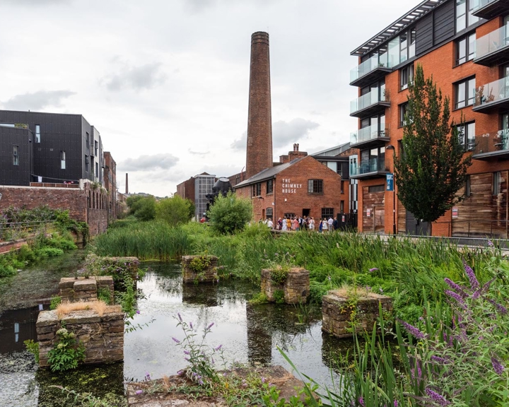 Building, a mixture of old and new, stand along the banks of a river. In the distance is a chimney stack rising up out of Kelham Island Museum.
