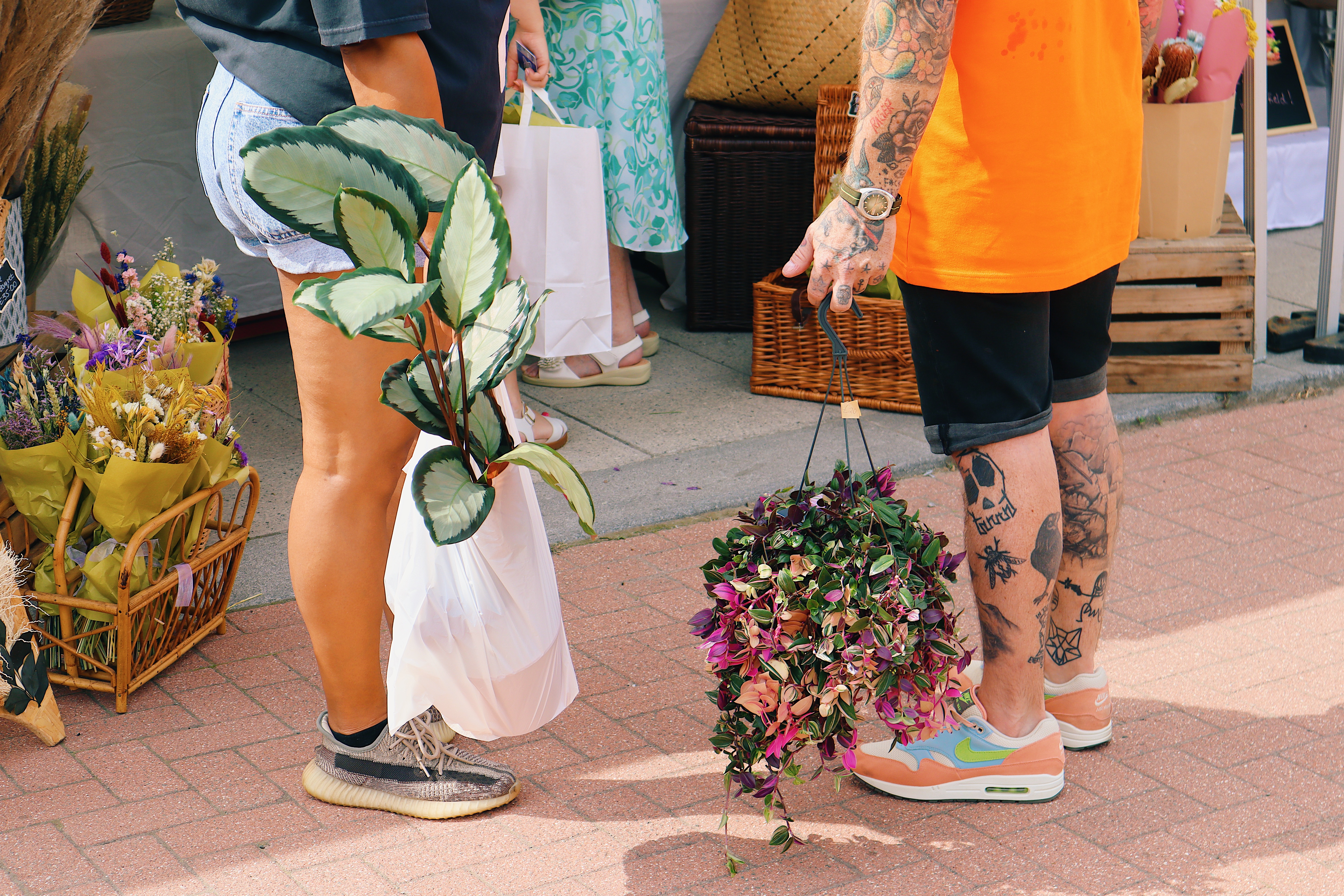 Two people standing on a paved walkway at an outdoor market, each holding plants. One person holds a leafy green plant in a white plastic bag, while the other carries a hanging basket of purple and green foliage. Behind them are baskets of dried flowers and wooden crates near a stall.