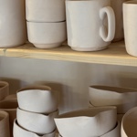 Stacks of unglazed off‑white ceramic mugs and bowls arranged on wooden shelves, showing smooth matte surfaces and simple handmade forms.