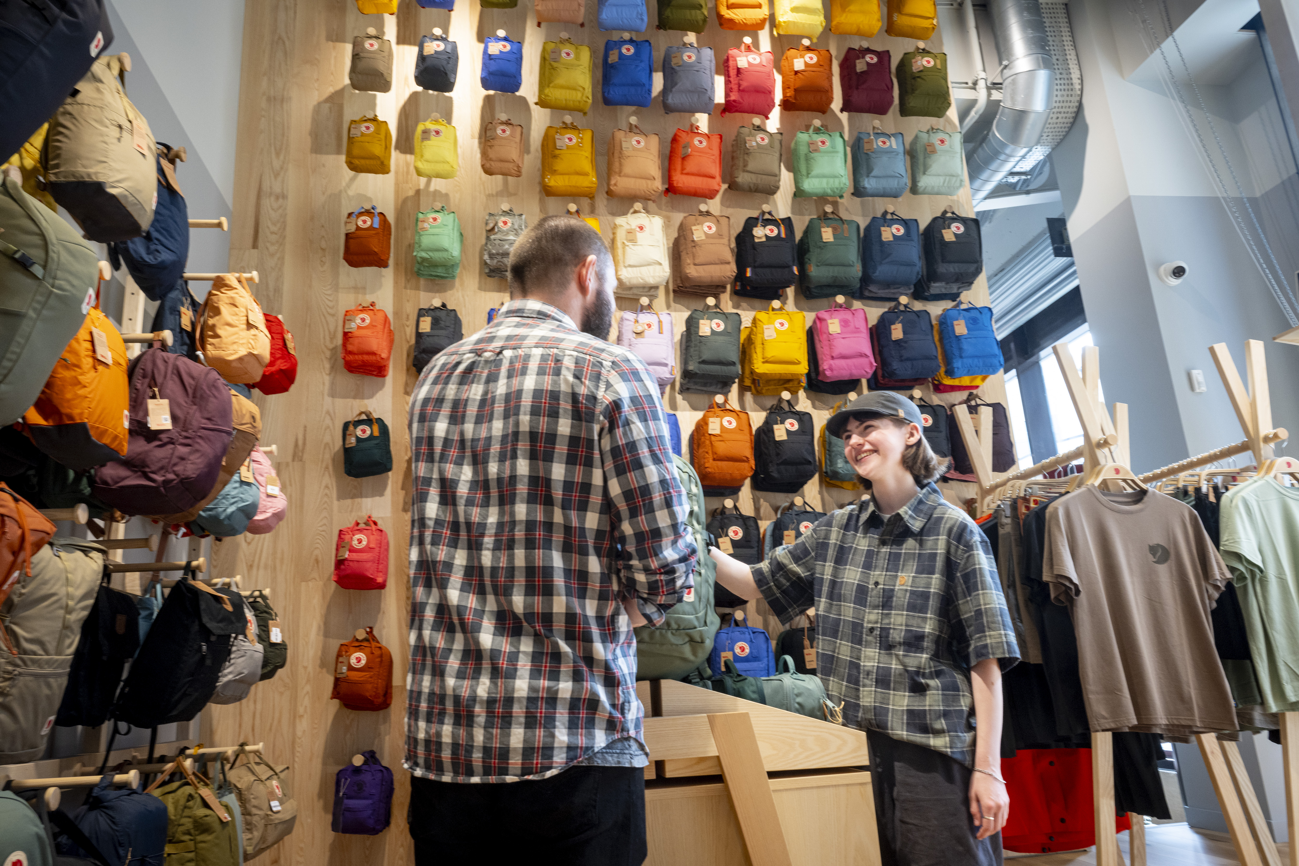 Inside a retail store with a wooden display wall covered in colorful backpacks arranged in neat rows. Two people stand near a light wooden counter, one appearing to assist the other. To the right, racks display neatly hung t-shirts in neutral tones. The store has bright lighting and a modern, organized layout.