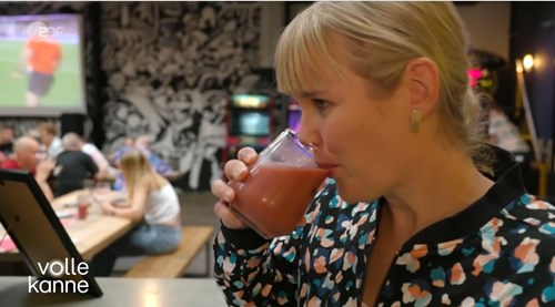“Person holding a glass of pink beverage inside a casual dining area with wooden tables and benches. Several people are seated in the background, and a large screen showing a sports game is visible on the wall along with black-and-white mural artwork. Text on the bottom left reads ‘volle kanne.’”