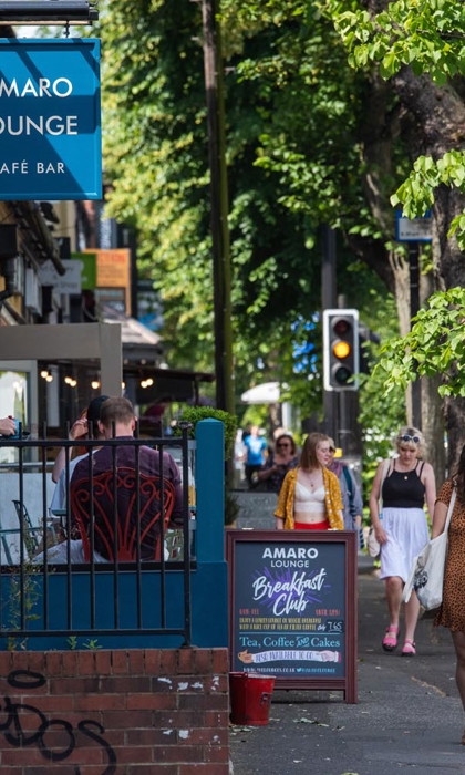 A row of shops and restaurants on a tree lined street. There are people walking up and down the pavement.