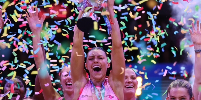 A netball team celebrates a championship win as a player lifts a silver trophy overhead. Colourful confetti fills the air, and teammates raise their arms in celebration, all wearing bright pink uniforms. The scene is energetic and festive, with vivid lighting highlighting the moment of victory.