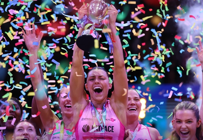 A netball team celebrates a championship win as a player lifts a silver trophy overhead. Colourful confetti fills the air, and teammates raise their arms in celebration, all wearing bright pink uniforms. The scene is energetic and festive, with vivid lighting highlighting the moment of victory.