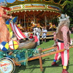 Two performers in Viking-themed costumes stand beside a decorated cart shaped like a wooden boat, complete with shields, a striped sail, and fishing nets. The cart is on grass in front of a brightly lit carousel with colourful horses. People in summer clothing watch the scene, and trees and a historic building are visible in the background, creating a lively festival atmosphere.