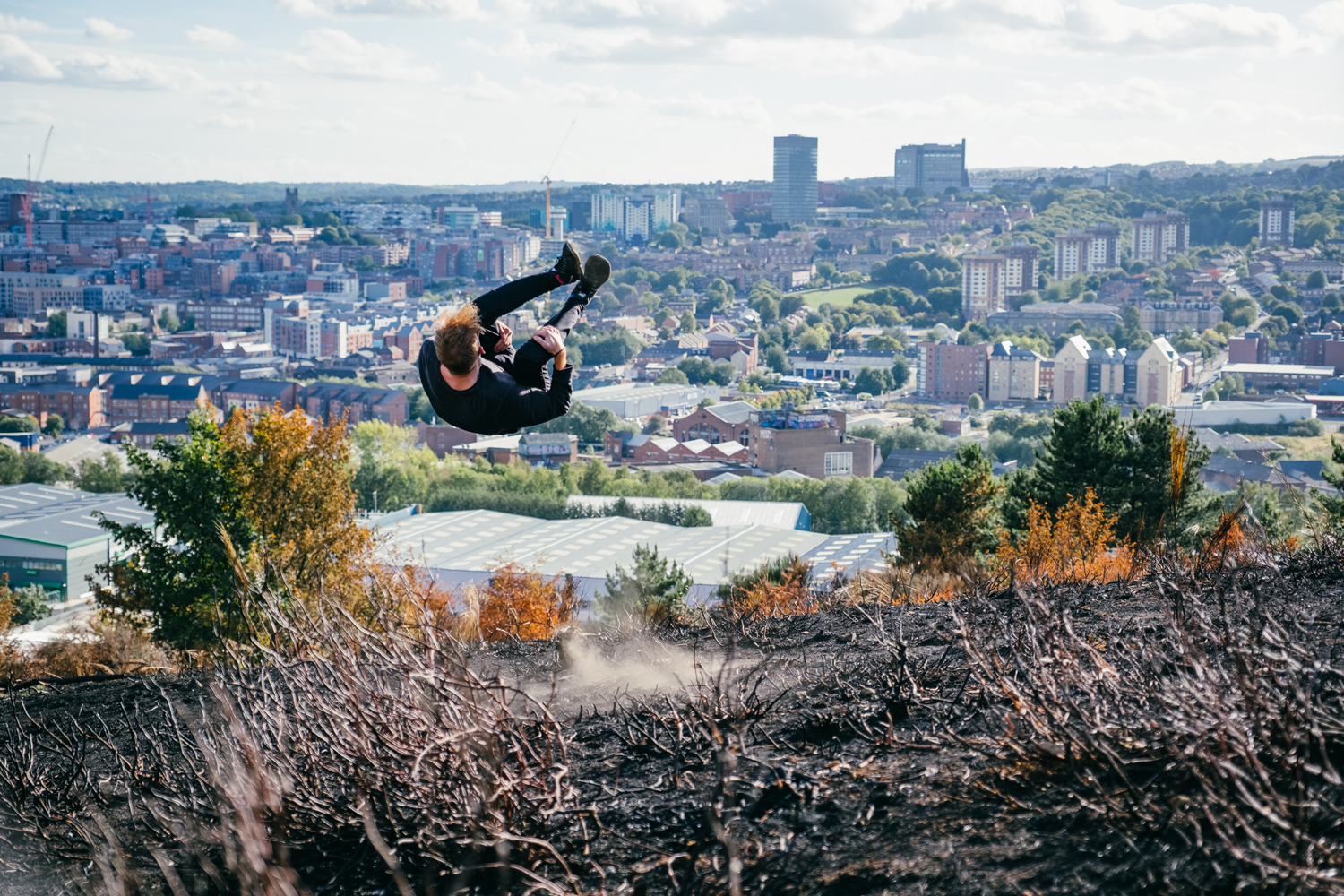 Person performing a mid-air flip on a hillside with burnt vegetation in the foreground, overlooking a cityscape with numerous buildings, trees, and distant hills under a partly cloudy sky.