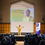 A speaker stands on a stage in a large hall, facing an audience seated in rows. The speaker is positioned in front of a large screen displaying the words “Lemn Sissay – let the light pour in live.” The hall has tall wooden paneling, pipe-organ–style architectural features, and stage lighting equipment on both sides. A banner to the right of the stage promotes the Off the Shelf festival. The audience fills the space, watching the presentation.