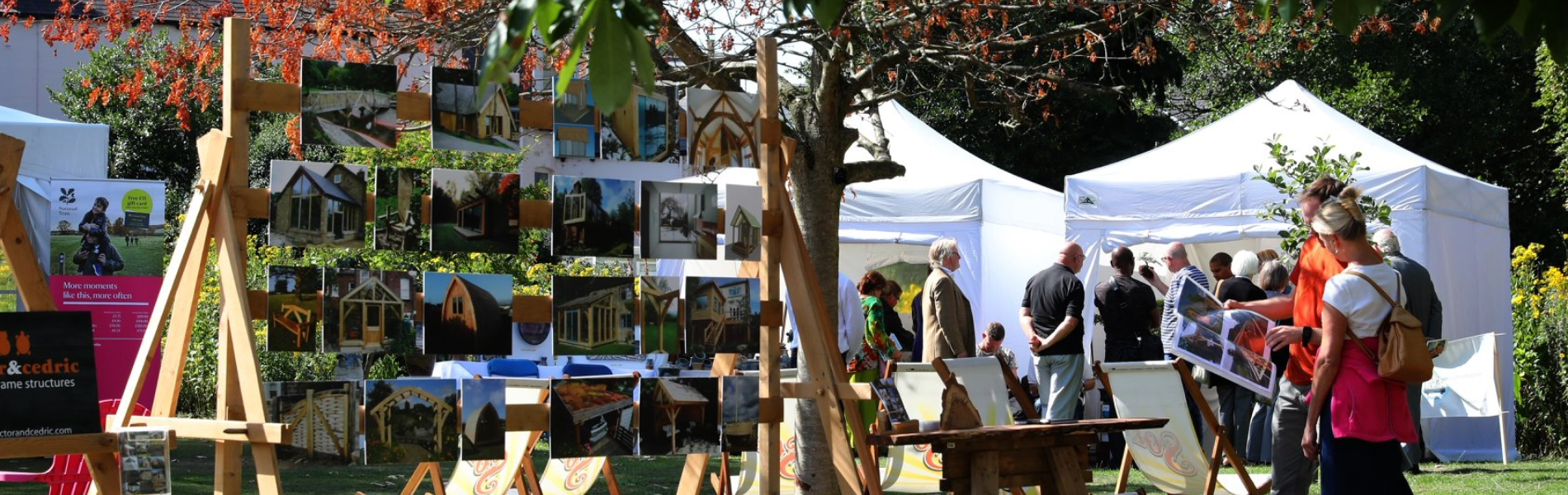 An outdoor art display at a garden event features rows of photographs or paintings mounted on wooden easels under a tree with autumnal leaves. Several deck chairs are arranged on the grass, and white marquee tents in the background host more visitors and exhibits. The scene is bright and sunny, with people browsing the artwork and enjoying the relaxed setting surrounded by greenery.