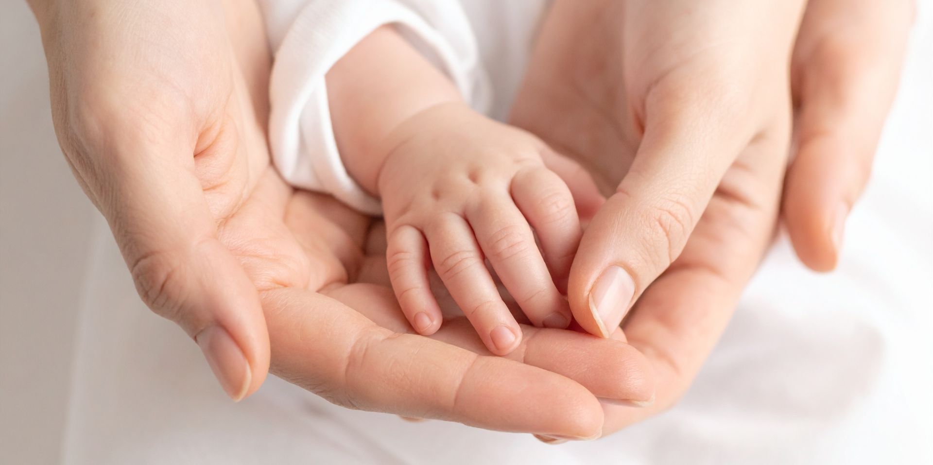 Close‑up of an adult gently holding a baby’s hand. The baby’s small fingers rest in the adult’s open palms, creating a soft, warm and nurturing scene against a light background.