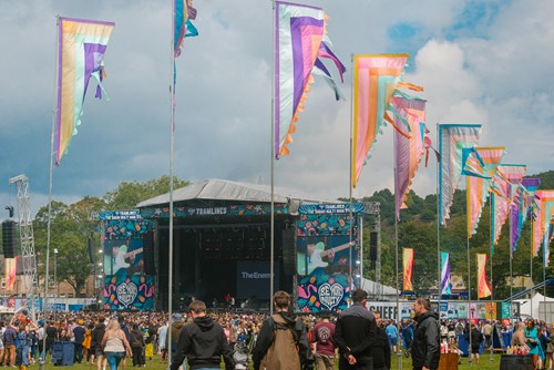 Outdoor music festival scene with a large stage displaying screens and banners reading ‘TRAMLINES.’ Colorful vertical flags line the area, and a crowd of people is gathered on the grassy field under a partly cloudy sky.