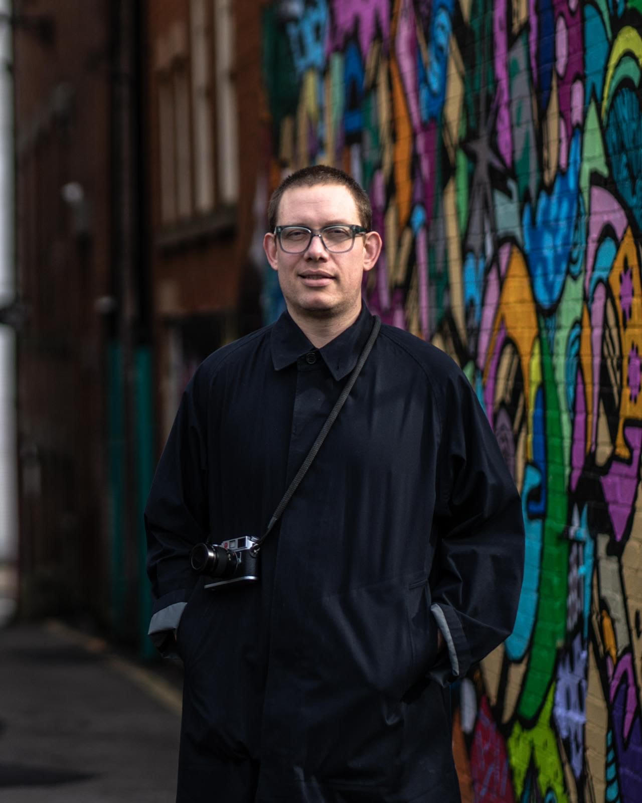 A man in a long, dark coat, with a camera slung over his shoulder, stands in front of some colourful street art.