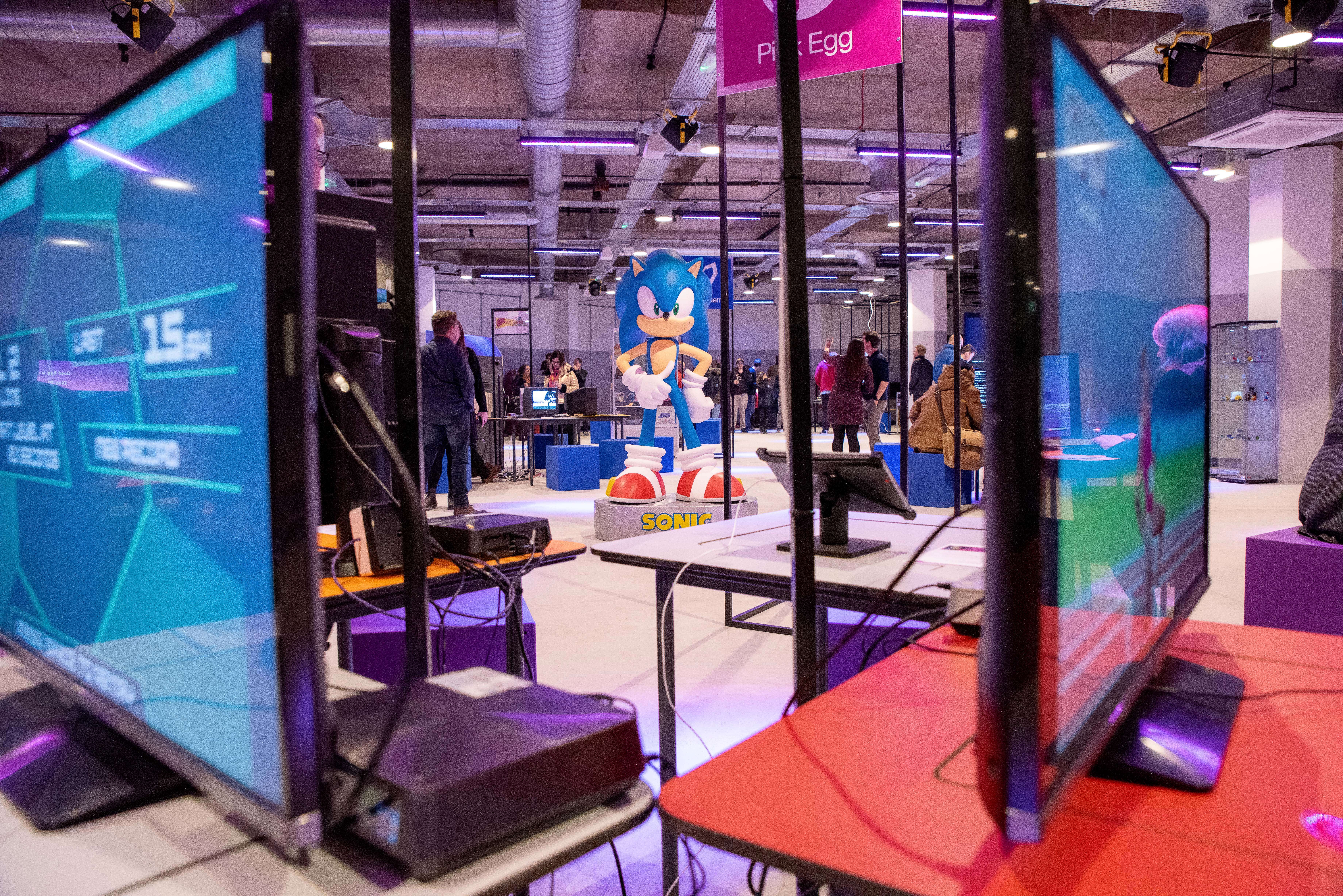 Interior of the National Videogame Museum with multiple gaming monitors in the foreground and a large Sonic the Hedgehog figure in the center. People are scattered around the space, interacting with exhibits. The room has an industrial ceiling with exposed pipes and bright lighting, creating a modern gaming environment.