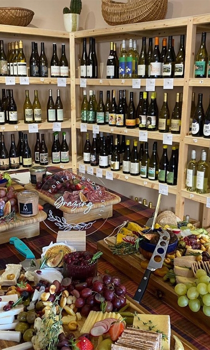 Rows of shelving filled with bottles of wine. In the centre of the room is a table covered in artfully arranged cheese, meats, grapes and other goods for sale.