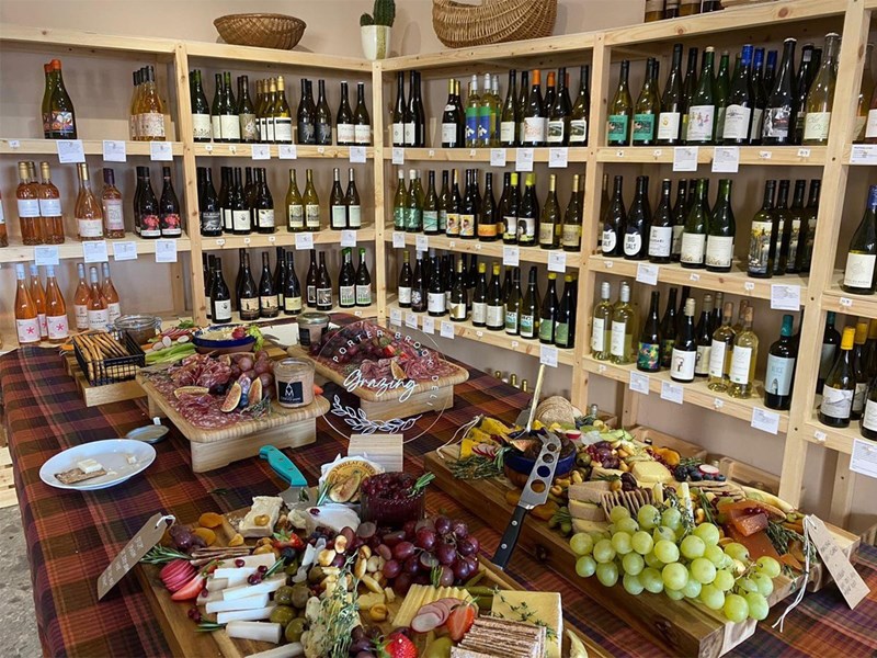 Rows of shelving filled with bottles of wine. In the centre of the room is a table covered in artfully arranged cheese, meats, grapes and other goods for sale.