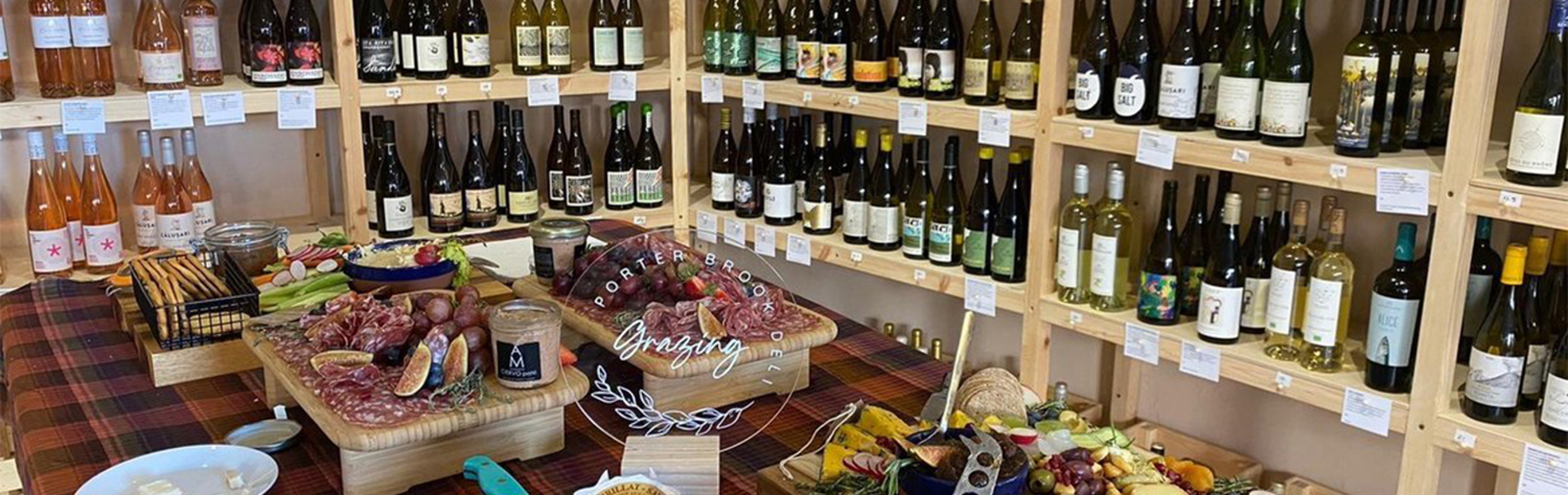 Rows of shelving filled with bottles of wine. In the centre of the room is a table covered in artfully arranged cheese, meats, grapes and other goods for sale.