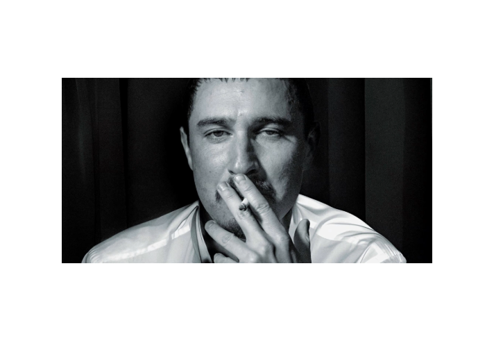 A person wearing a light-colored, long-sleeved shirt is seated against a dark background with vertical panels. One hand is raised near the chin, partially covering the mouth, creating a contemplative pose. The lighting emphasises the folds and sheen of the shirt, contrasting with the shadowed backdrop.