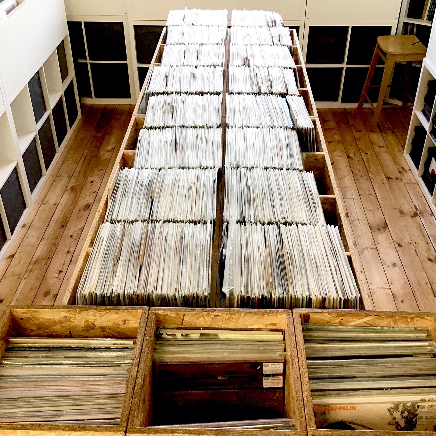 A wooden-floored room with long wooden tables filled with rows of vinyl records organized in sections. The records are stored vertically in wooden crates, and the tables run down the center of the room. Shelving units line the walls, and a wooden stool is visible on the right side.