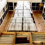 A wooden-floored room with long wooden tables filled with rows of vinyl records organized in sections. The records are stored vertically in wooden crates, and the tables run down the center of the room. Shelving units line the walls, and a wooden stool is visible on the right side.