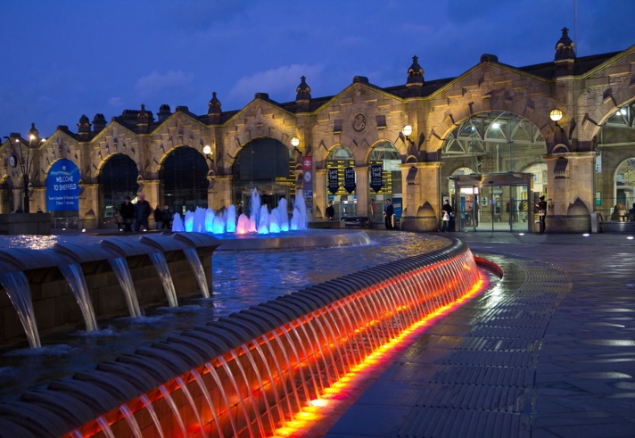 A nighttime view of Sheffield railway station featuring its historic stone façade with arched windows and illuminated signage. In the foreground, a curved water feature with cascading streams is lit by vibrant red and orange lights, while fountains in the background glow in shades of blue and purple. The scene is set against a deep blue sky, creating a striking contrast with the warm architectural lighting.