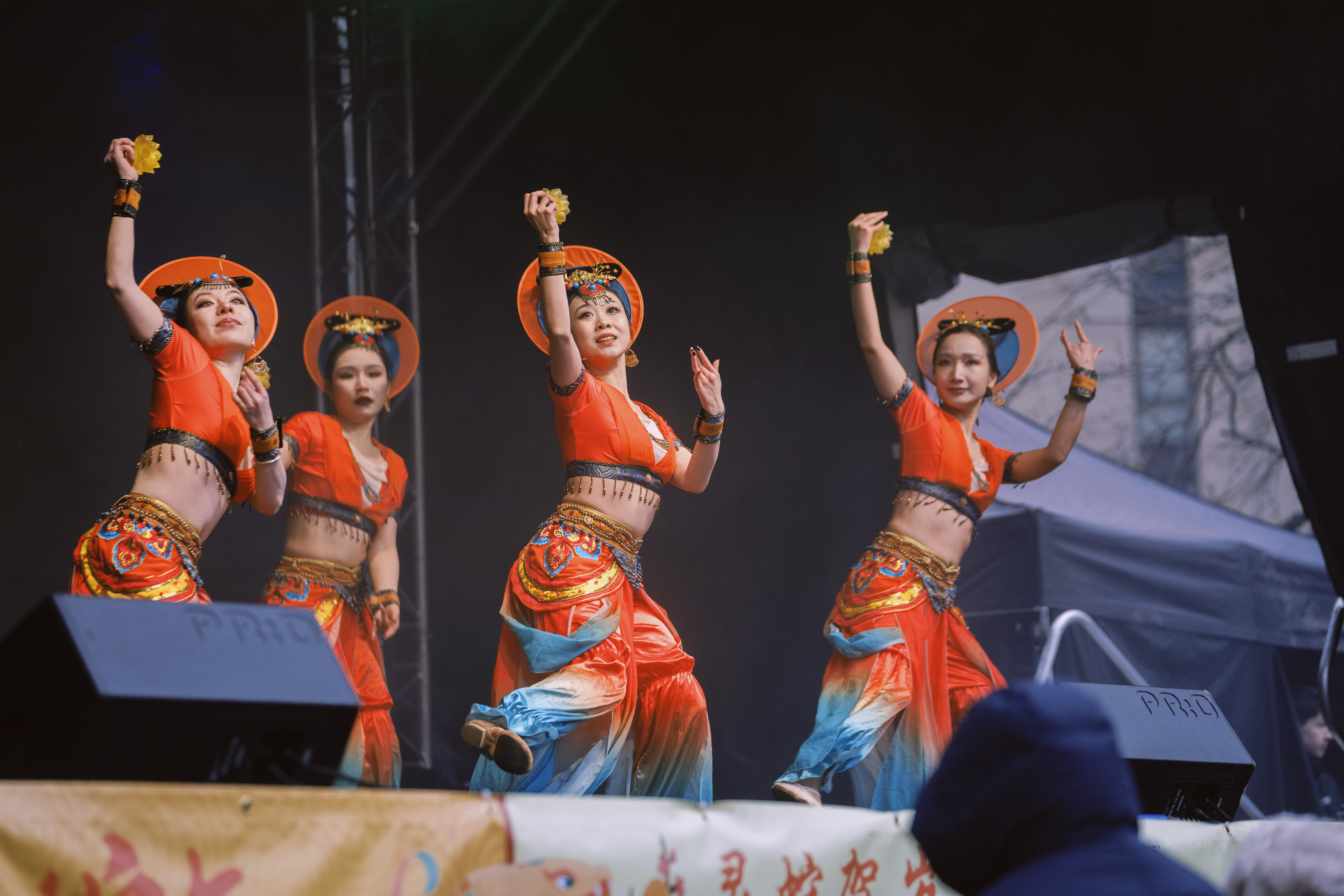 A group of performers on stage wearing coordinated orange and blue costumes with wide-brimmed hats, captured mid-dance with arms raised. The stage is decorated with a banner featuring Chinese characters.