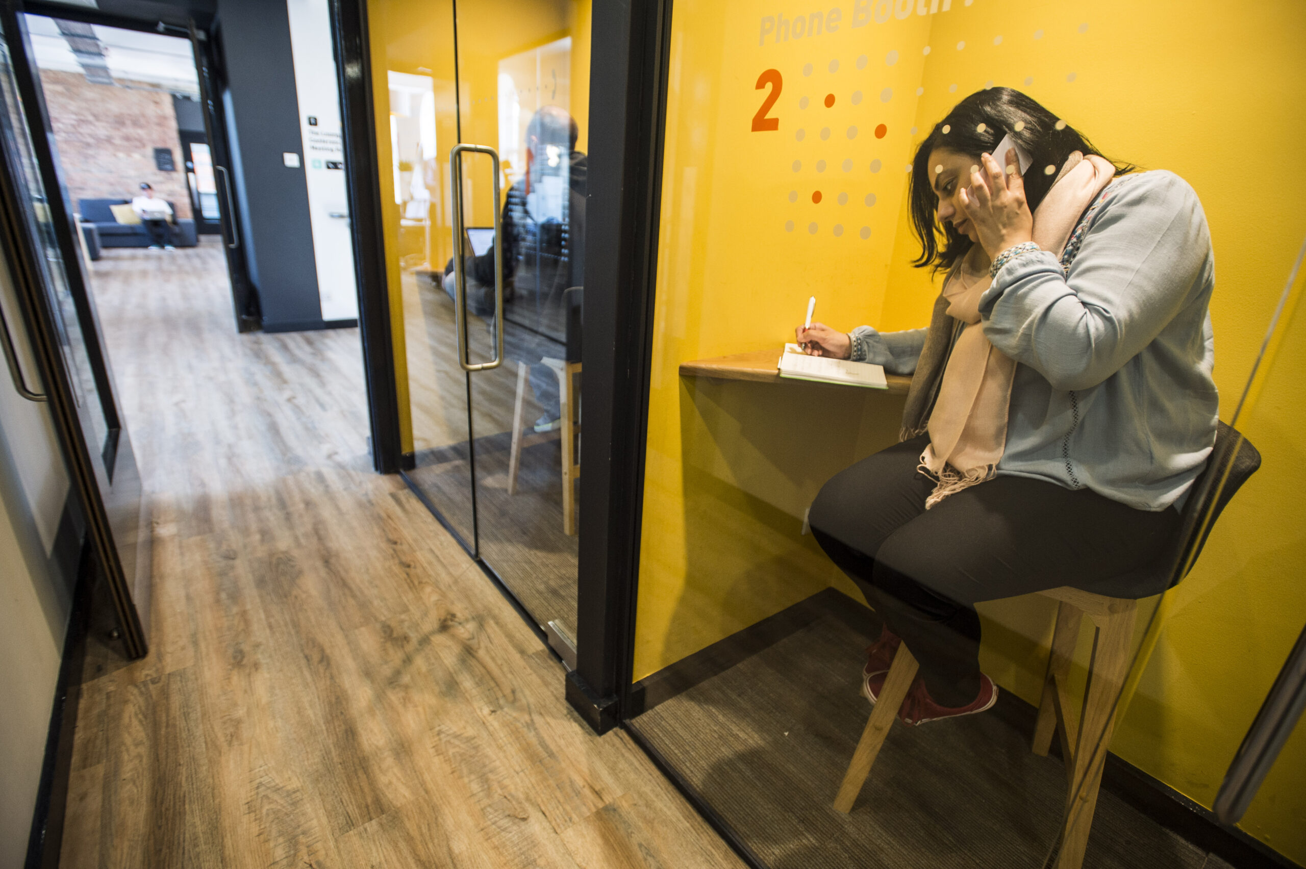 A woman sat on a stool at a desk talking on her mobile phone.