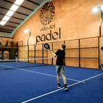 Indoor padel court with blue flooring, where two players prepare to rally while others watch from a raised viewing area against an orange wall displaying the ‘Vida del Padel’ logo