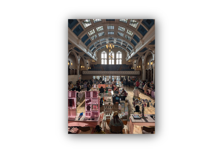 Indoor craft market in a grand hall with a high arched ceiling, large windows, and hanging lights. Several stalls with colourful displays of prints, jewellery, and handmade items line the wooden floor, with people browsing and sitting behind tables. Pink shelving units and signs like “Prints” and “Keychains” are visible in the foreground. Balcony seating and ornate architectural details frame the space.