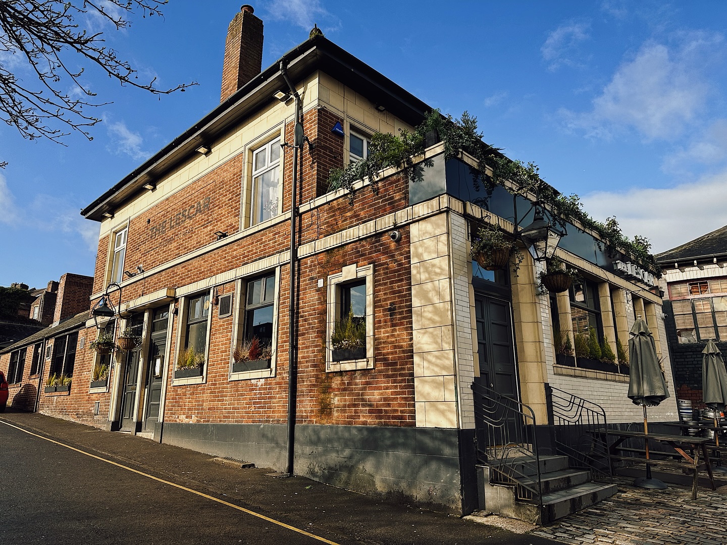 The exterior of The Lescar pub, a traditional brick building with cream detailing, large windows and hanging plants, photographed on a bright day.