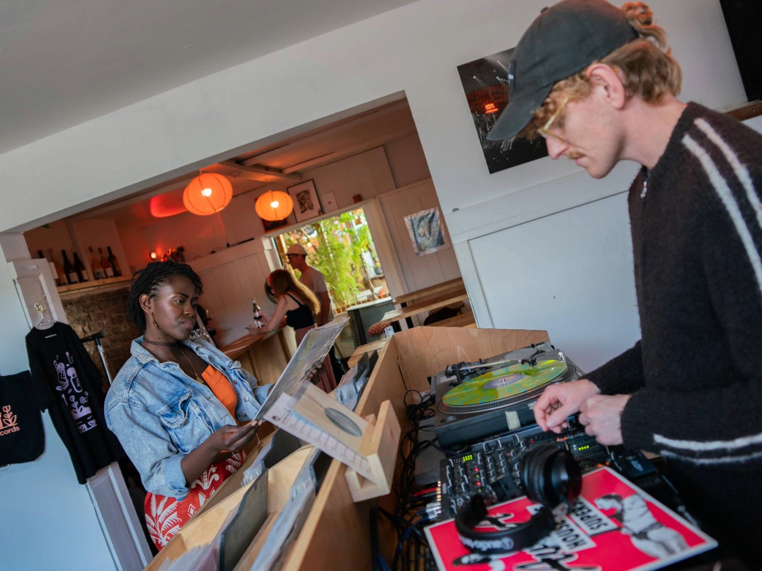 A person stands at a wooden record counter browsing vinyl records, while another operates DJ equipment with turntables and a mixer. Headphones and a colourful record sleeve are visible on the counter. The setting appears to be a cosy shop or café with white walls, hanging orange lights, shelves with bottles, and a tote bag displayed on the wall. A doorway in the background reveals more people and greenery outside.