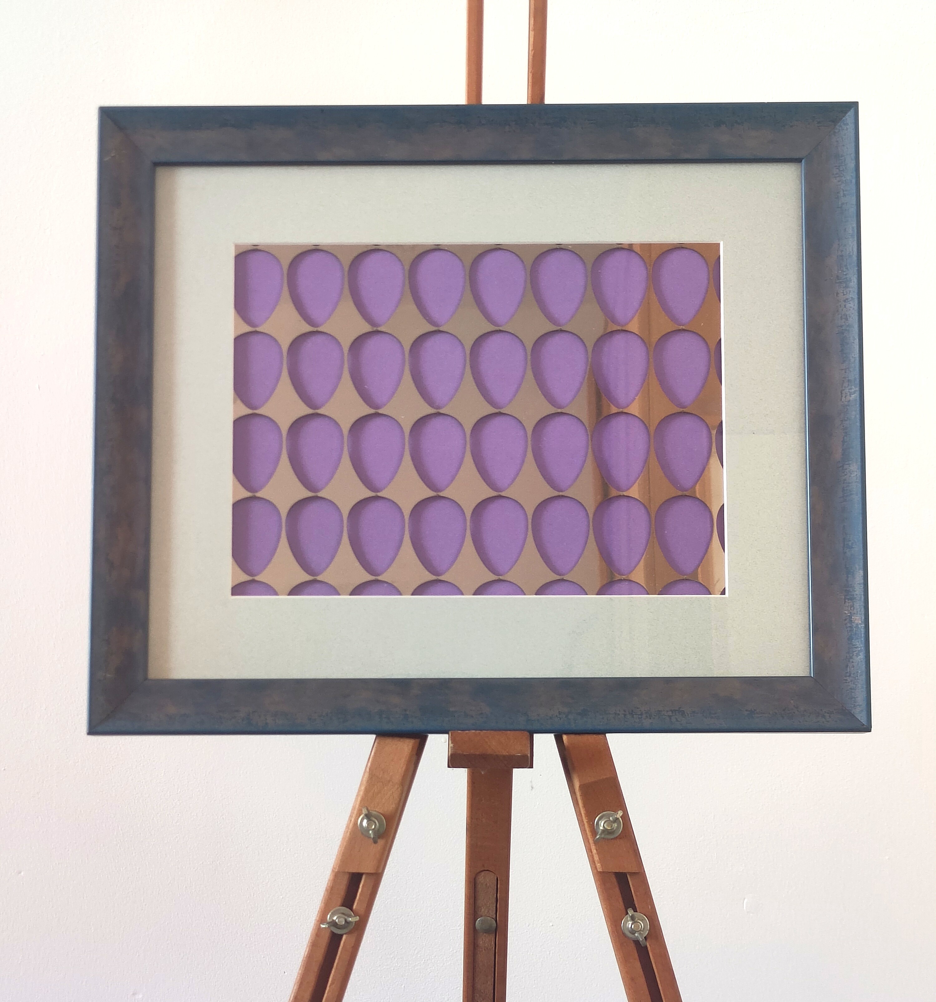 Framed artwork with a rustic wooden frame featuring a grid of purple teardrop shapes on a metallic background, displayed on an easel against a neutral wall.