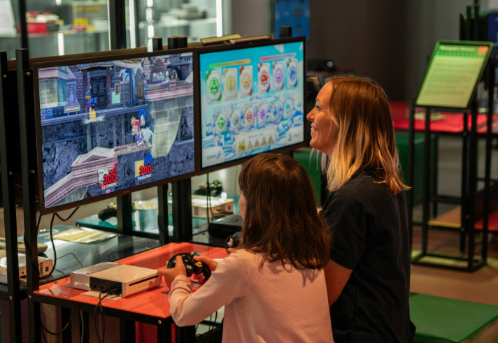 A person with a child sat next to them are playing a video game together. In the background are displays of video games from the history of the video game industry.
