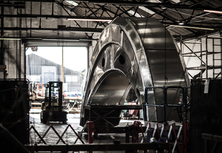 Large metallic structure resembling a curved boat hull under construction inside an industrial workshop. The piece is shiny and partially assembled, with scaffolding and barriers around it. A forklift and open workshop doors are visible in the background, along with steel beams and equipment.