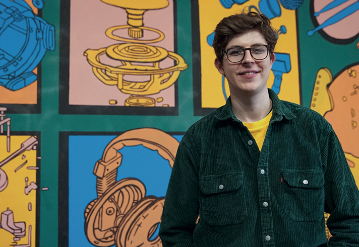 A person wearing a dark green button-up shirt over a yellow top is standing in front of a colorful mural featuring illustrated mechanical components, such as gears, headphones, and engine parts. The artwork is arranged in panels with vibrant shades of blue, orange, and green, creating a bold and technical-themed background.