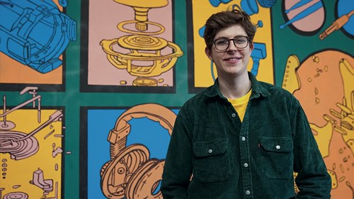 A person wearing a dark green button-up shirt over a yellow top is standing in front of a colorful mural featuring illustrated mechanical components, such as gears, headphones, and engine parts. The artwork is arranged in panels with vibrant shades of blue, orange, and green, creating a bold and technical-themed background.
