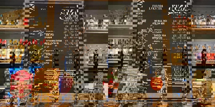 The bar at Guzzle with several beer pumps, a large blackboard with drinks and prices and shelves filled with bottles.