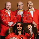 A picture of the band which consists of 4 individuals, who are all wearing read blazers. One is sat on a chair in a laid back manner, and wearing sunglasses, and the rest are stood behind him. All looking at the camera