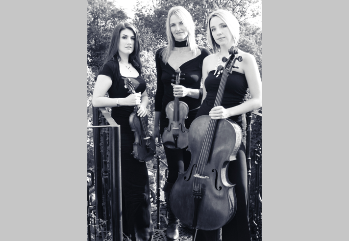 Black and white promo shot of the trio Simply Strings in an outdoor setting. One holds a cello and the other two hold violins.