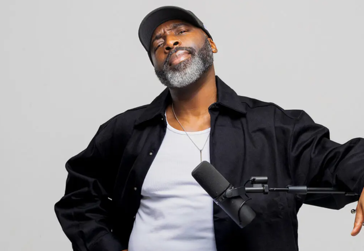 Person wearing a black jacket over a white shirt and a necklace, standing in front of a plain light gray background. The person is holding one arm out to the side and is positioned near a black microphone mounted on a boom arm. A dark cap is worn on the head.