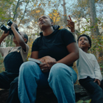 An adult and two children sit on a log in a forest. One of the children is looking through a pair of binoculars. 