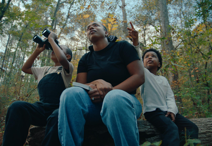 An adult and two children sit on a log in a forest. One of the children is looking through a pair of binoculars. 